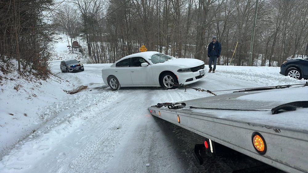 A white Dodge Charger winched onto the Top Notch flatbed on a snow-covered road, a sheriff's vehicle on scene